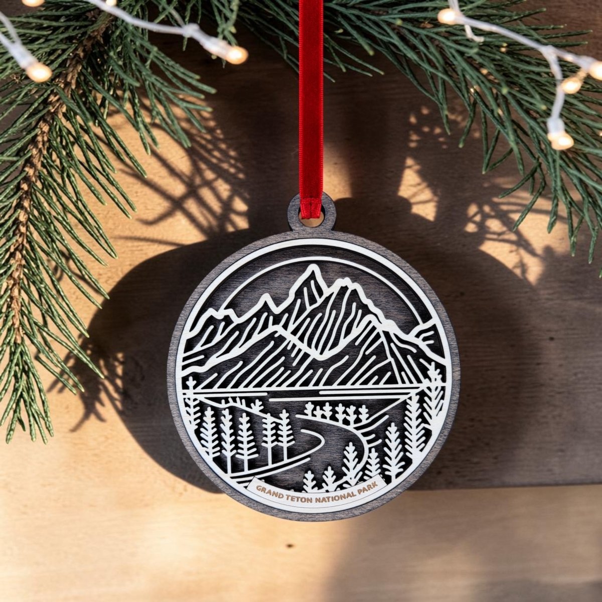 The Grand Teton National Park Line Art Ornament by Mirabilia Boutique features an oval cutout design showcasing mountains, trees, and a winding river. It hangs from a red ribbon with pine branches and string lights in the background.