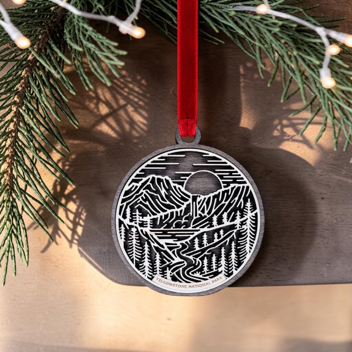 The Yellowstone National Park Line Art Ornament by Mirabilia Boutique is an oval wooden keepsake featuring a beautifully detailed nature scene with mountains, trees, a river, and the sun. Hanging from a red ribbon on a fir branch, this ornament showcases elegant line art and the words "Yellowstone National Park.