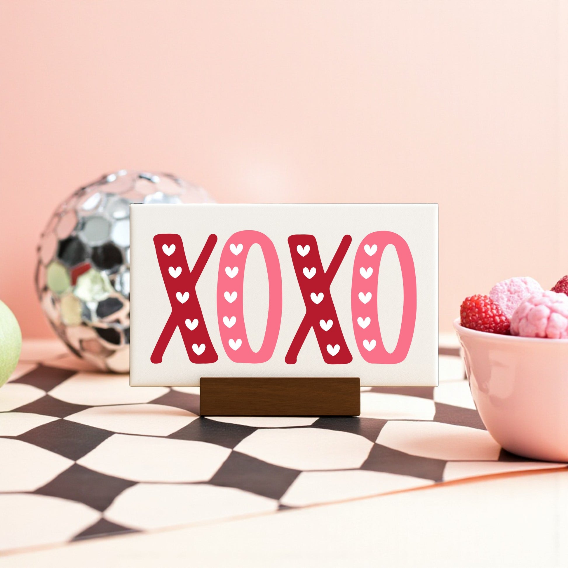 The XOXO Valentine's Sign by Mirabilia Boutique features red and pink hearts on a 6x4 canvas with a wood base—ideal Valentine’s Day decor—displayed beside colorful candies and a disco ball on a geometric-patterned surface.