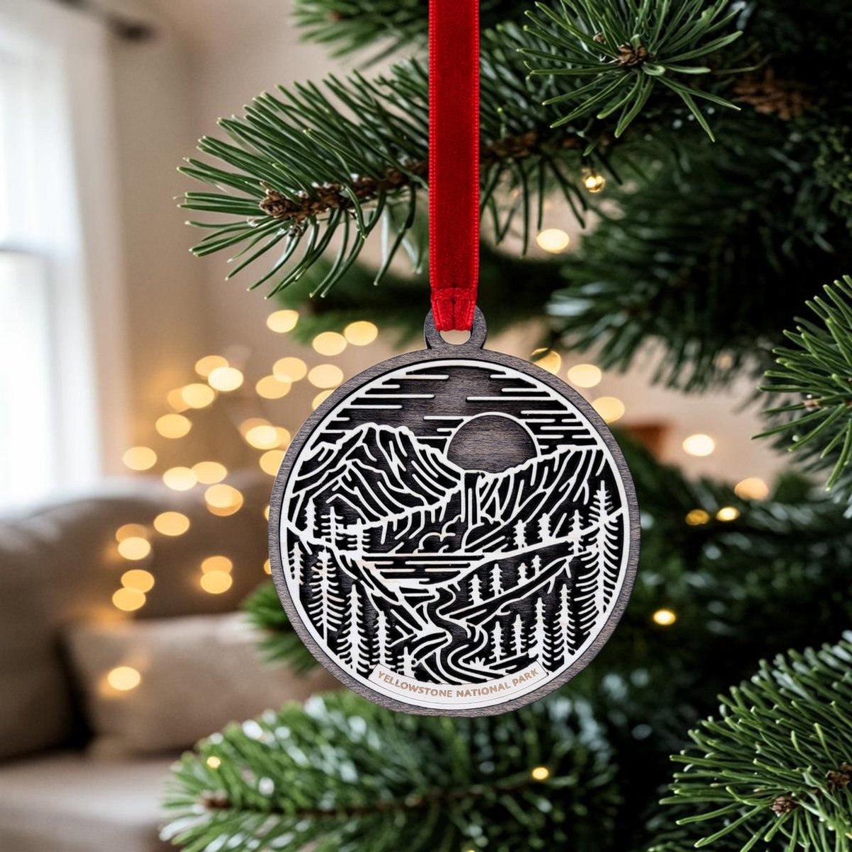 A Mirabilia Boutique Yellowstone National Park Line Art Ornament, featuring an oval shape with a mountain and river design, hangs on a Christmas tree by a red ribbon; blurred lights and a couch appear in the background.