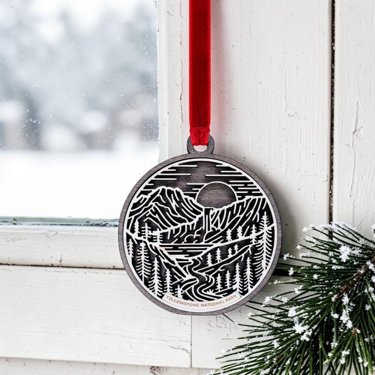 The Yellowstone National Park Line Art Ornament by Mirabilia Boutique features a detailed landscape design and hangs from a red ribbon on a white window frame, surrounded by snowy evergreen branches and a wintry scene outside. This ornament beautifully captures the essence of Yellowstone’s natural beauty, making it a unique and memorable keepsake for any holiday display.