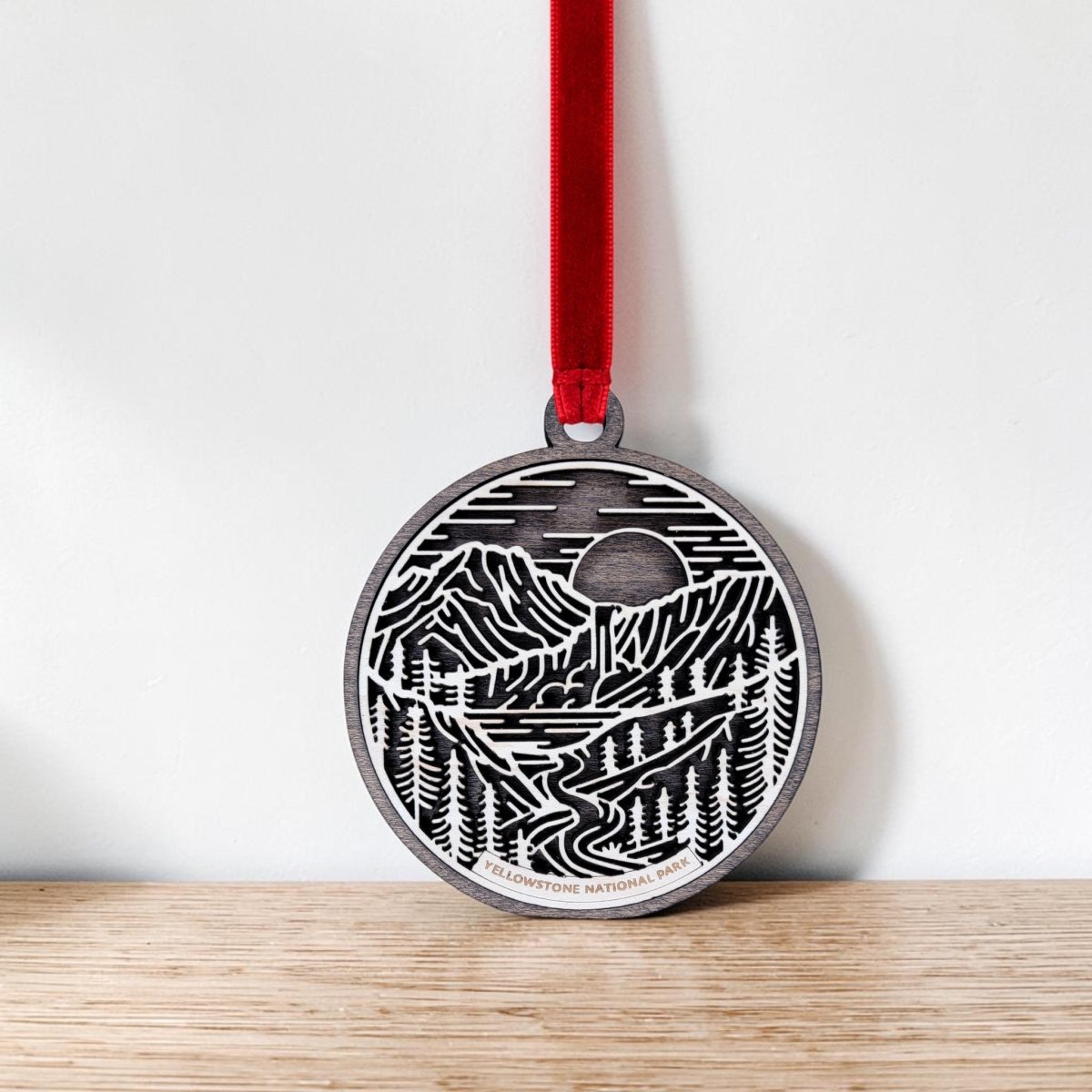 The Yellowstone National Park Line Art Ornament by Mirabilia Boutique features a mountain, trees, river, and sun design in delicate line art. Hanging from a red ribbon, "Yellowstone National Park" is displayed at the bottom. The ornament is shown resting against a light wall on a wooden surface.