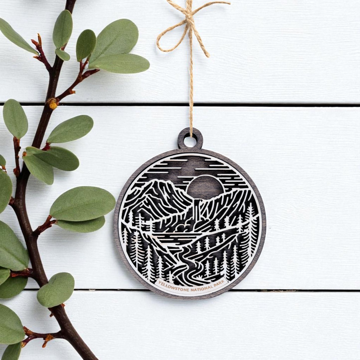 The Yellowstone National Park Ornament by Mirabilia Boutique is a round, black and white wooden piece featuring mountains, trees, and a river—handcrafted and hanging from twine—ideal as rustic holiday decor or a unique nature gift.