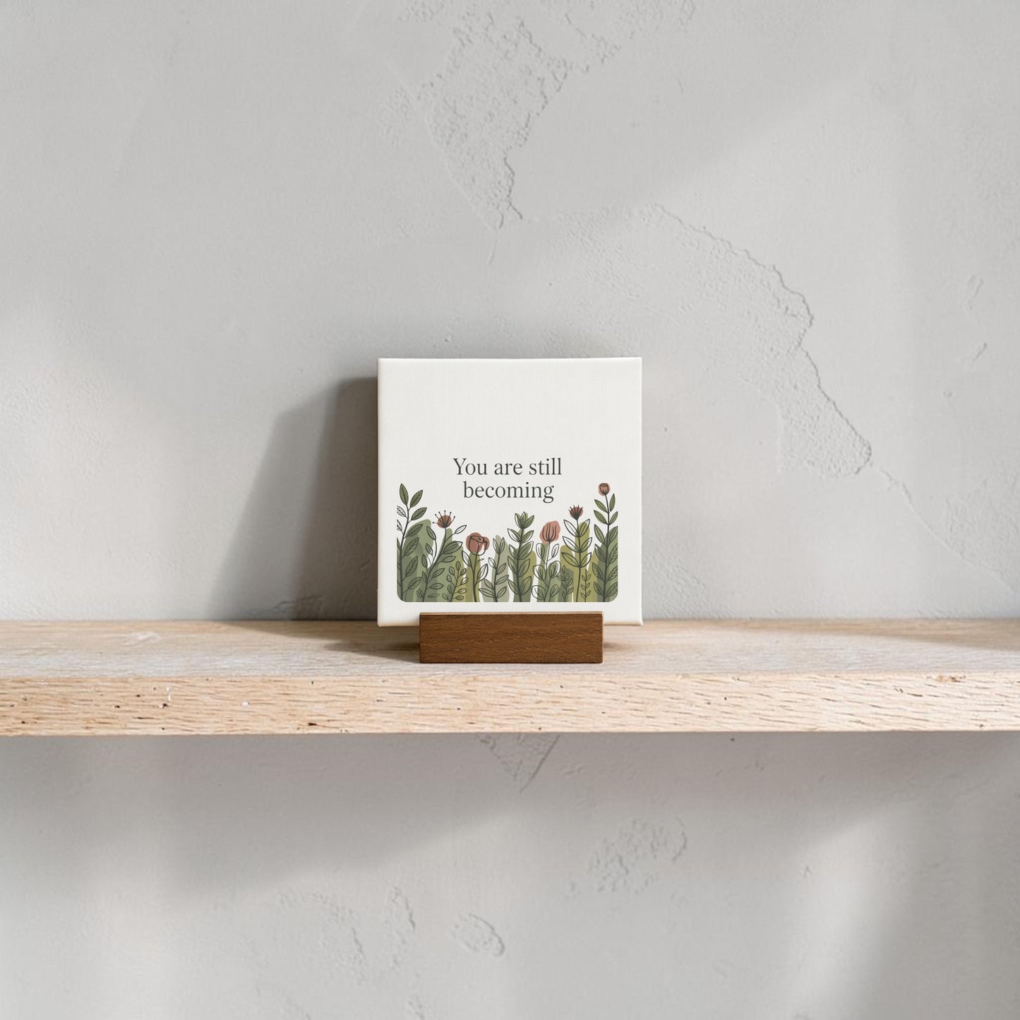 The Mirabilia Boutique "You Are Still Becoming" Mini Canvas Sign features floral art and an inspiring message on a wooden stand—ideal for botanical decor lovers, and stylishly displayed on a shelf against a gray textured wall.