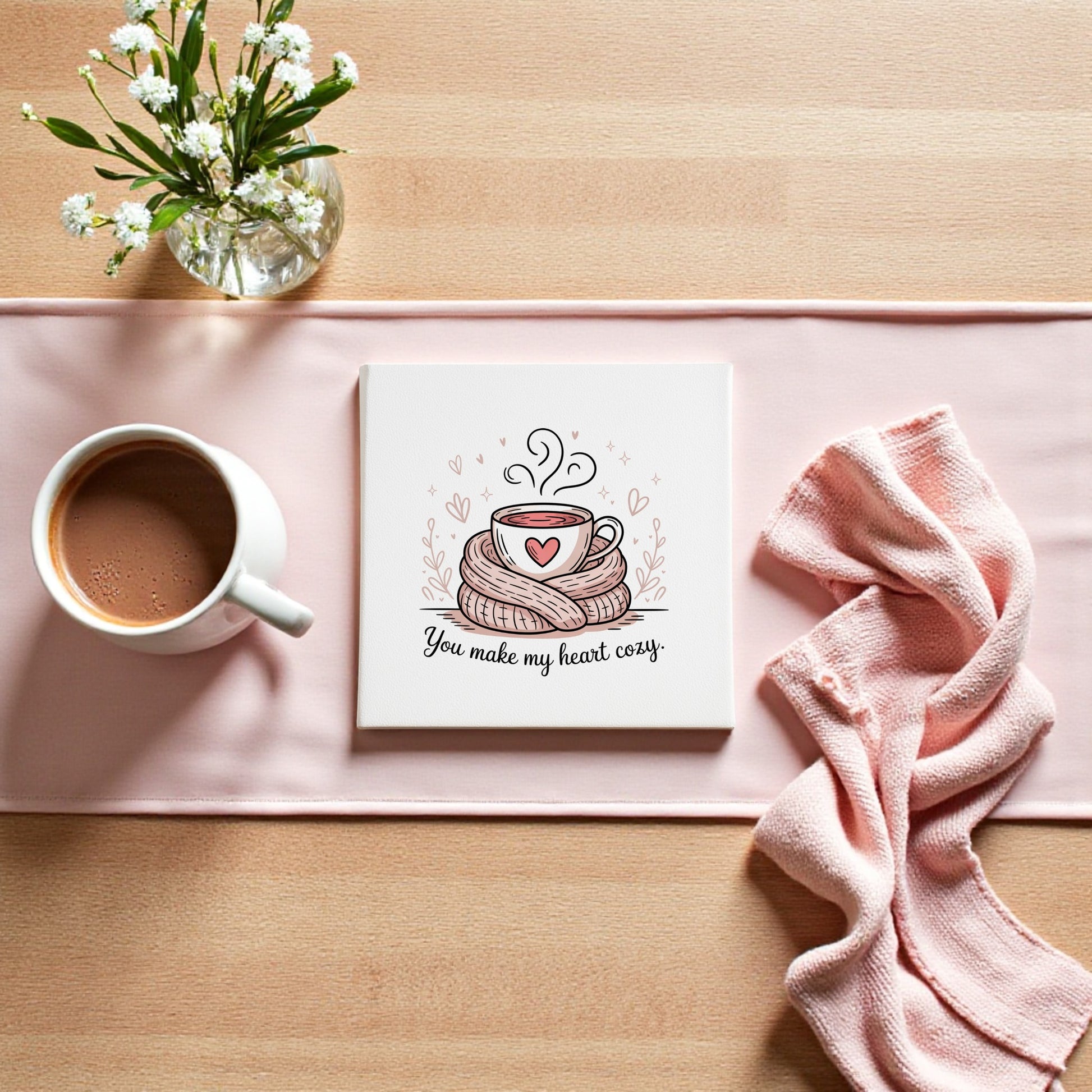A cup of hot chocolate rests beside a pink cloth, white flowers, and Mirabilia Boutique’s "You Make My Heart Cozy Canvas"—a romantic décor piece with a steaming mug illustration, ideal as a warm Valentine’s gift.