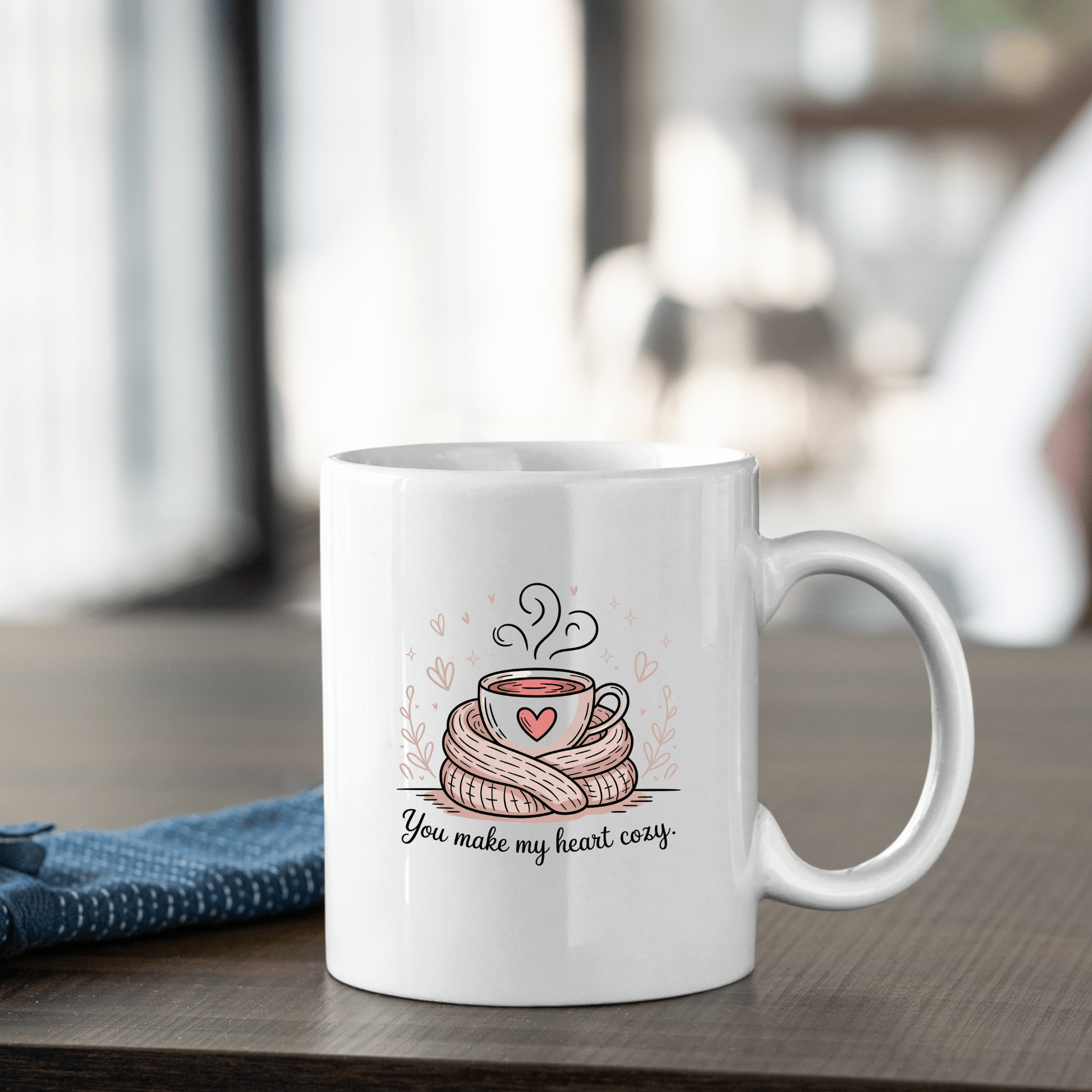 The You Make My Heart Cozy Mug by Mirabilia Boutique sits on a wooden table, featuring a steaming cup wrapped in a scarf with a heart and the text "You make my heart cozy," making it an ideal Valentine’s Day gift for coffee lovers.