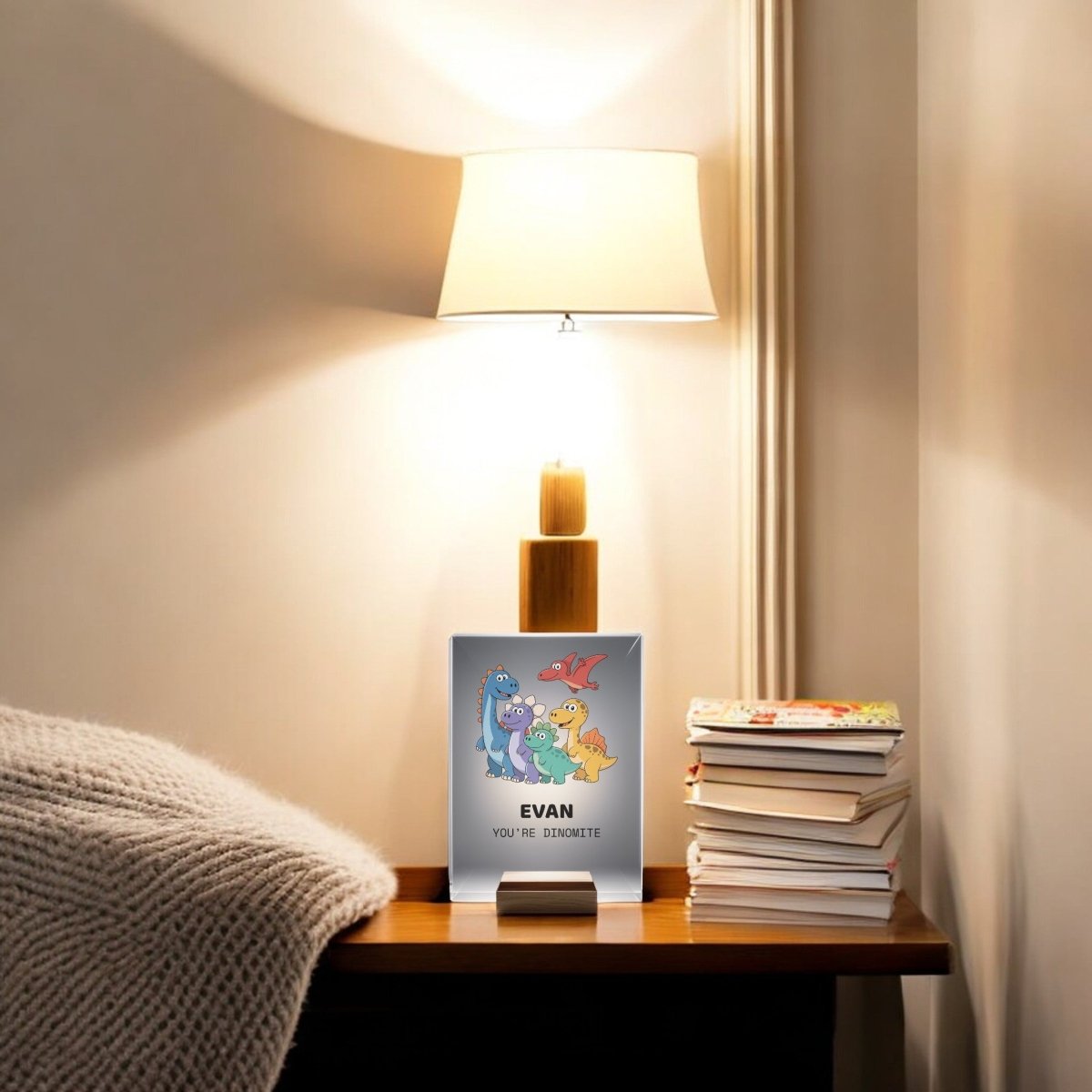 The Personalized Dinosaur Night Light for Kids by Mirabilia Boutique sits on a bedside table with books and a “EVAN YOU’RE DINO-MITE” card featuring dinosaurs—a fun, cozy touch for any child’s bedroom. The softly lit wall sets the mood.