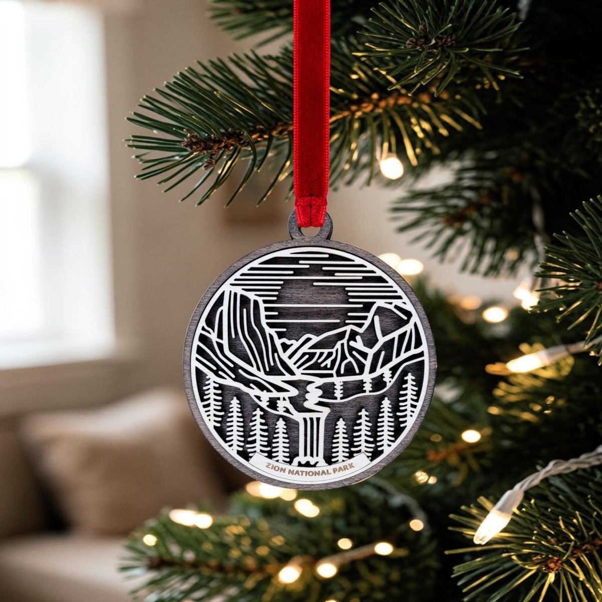 The Zion National Park Line Art Ornament by Mirabilia Boutique, crafted from metal and showcasing an intricate line art design of the park’s landscape, hangs from a red ribbon on a Christmas tree adorned with lights and set against a softly blurred background.