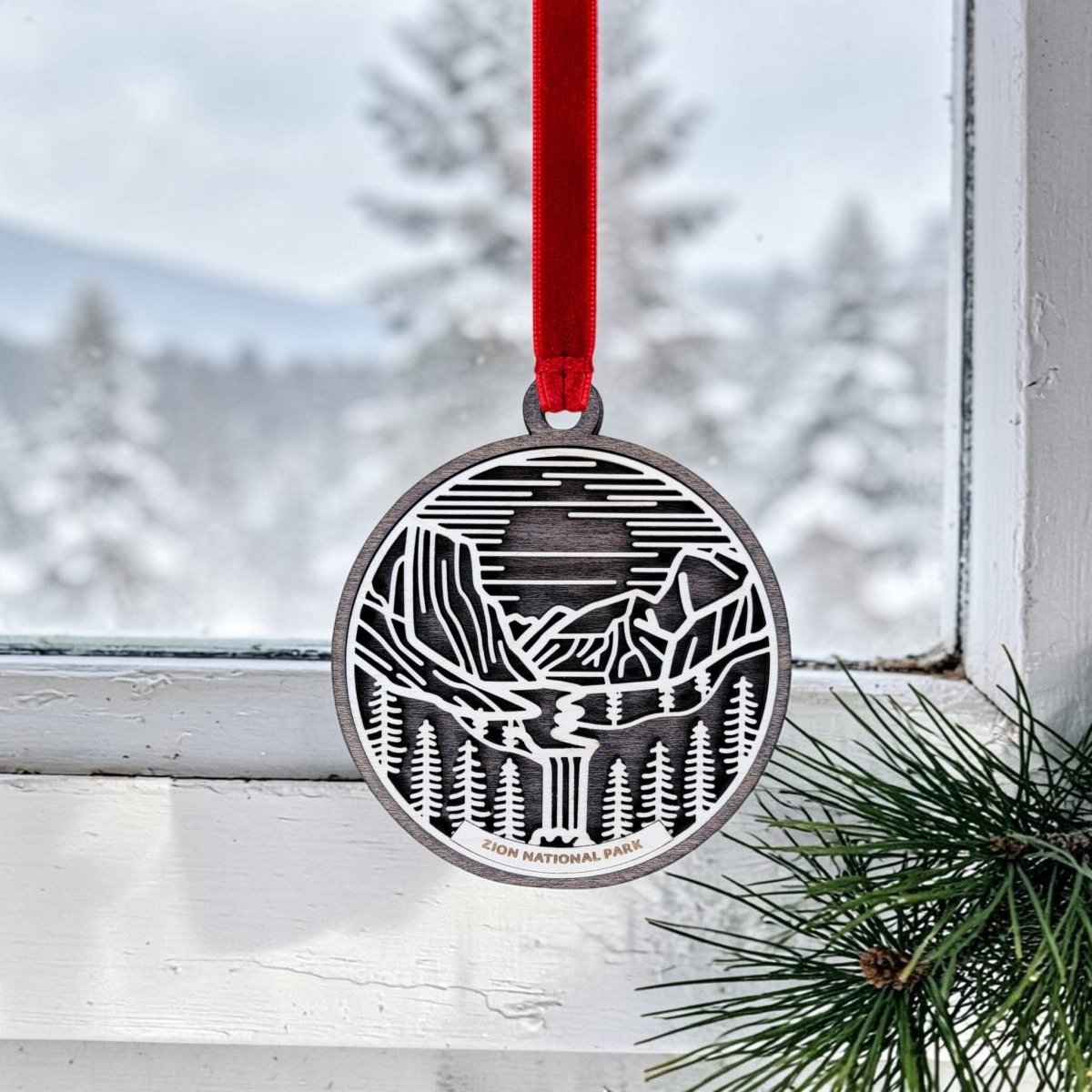 The Zion National Park Line Art Ornament by Mirabilia Boutique is a round wooden keepsake featuring a cutout scene of mountains, trees, and a river. Hanging by a red ribbon in a window, it’s complemented by the snowy trees and mountains outside, while a pine branch in the corner enhances the charm of its minimalist line art design.