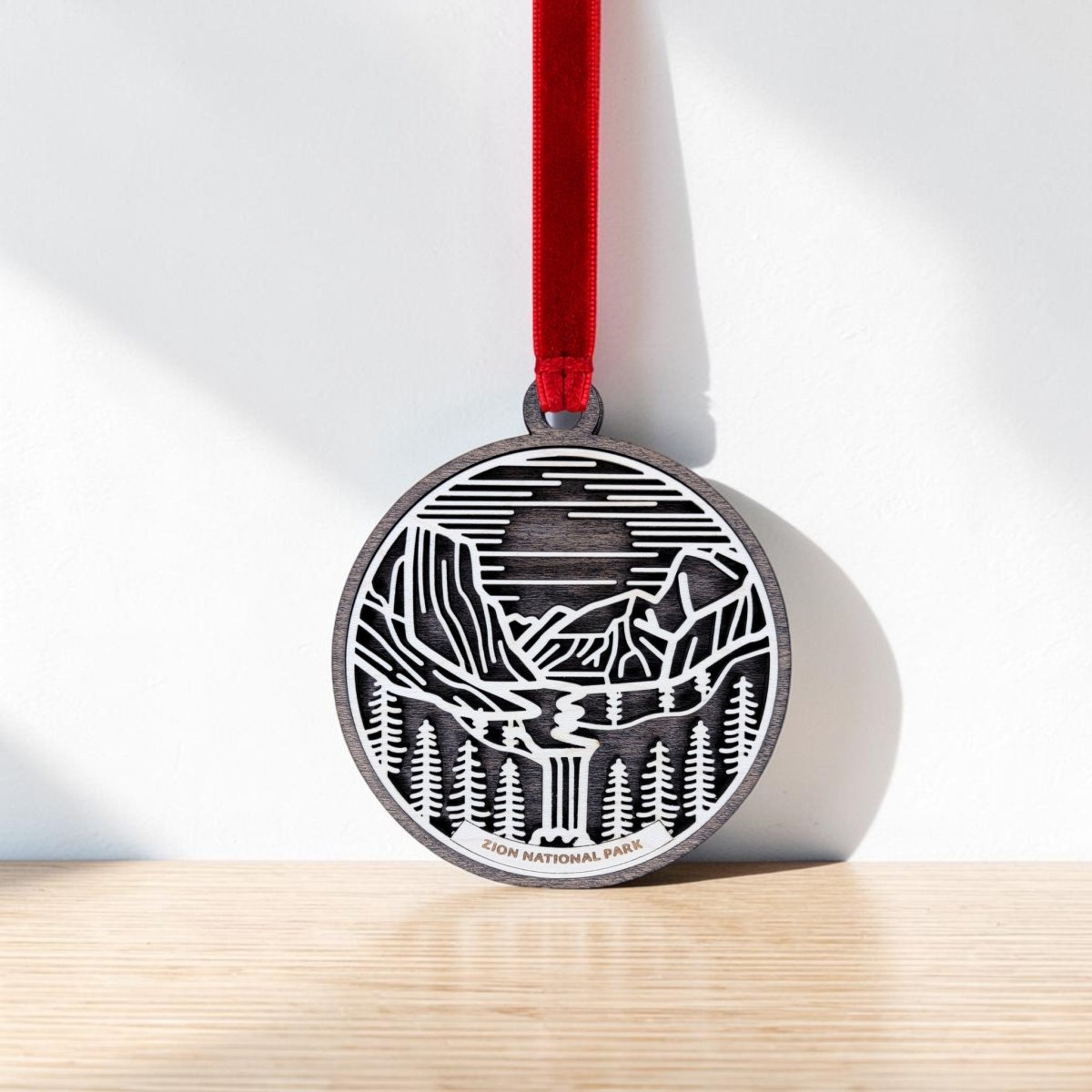 The Zion National Park Line Art Ornament by Mirabilia Boutique is a round metal piece featuring a striking black and white line art design of a mountain, river, and forest. It hangs from a red ribbon and has "Zion National Park" engraved at the bottom.