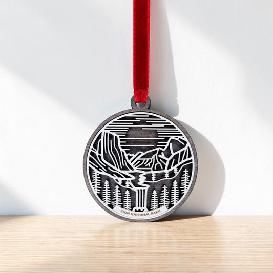 The Zion National Park Line Art Ornament by Mirabilia Boutique is a round metal piece featuring a striking black and white line art design of a mountain, river, and forest. It hangs from a red ribbon and has "Zion National Park" engraved at the bottom.