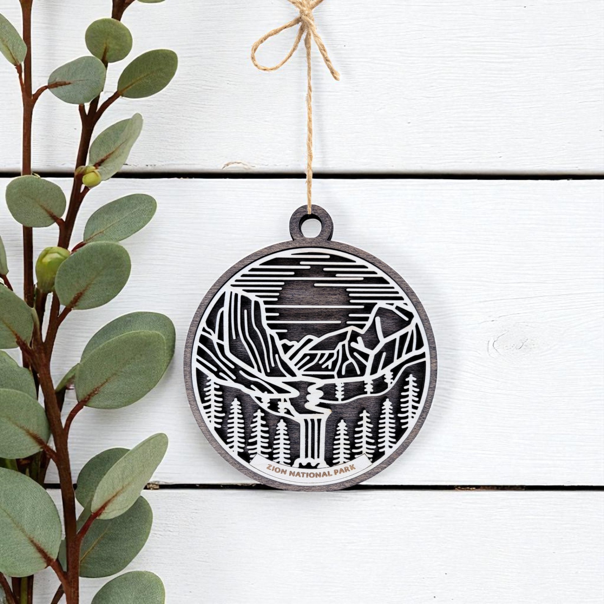 The Zion National Park Ornament by Mirabilia Boutique is a round wooden keepsake featuring cutout mountains, trees, and a river. It hangs from twine, making it a perfect handcrafted National Park gift or souvenir.